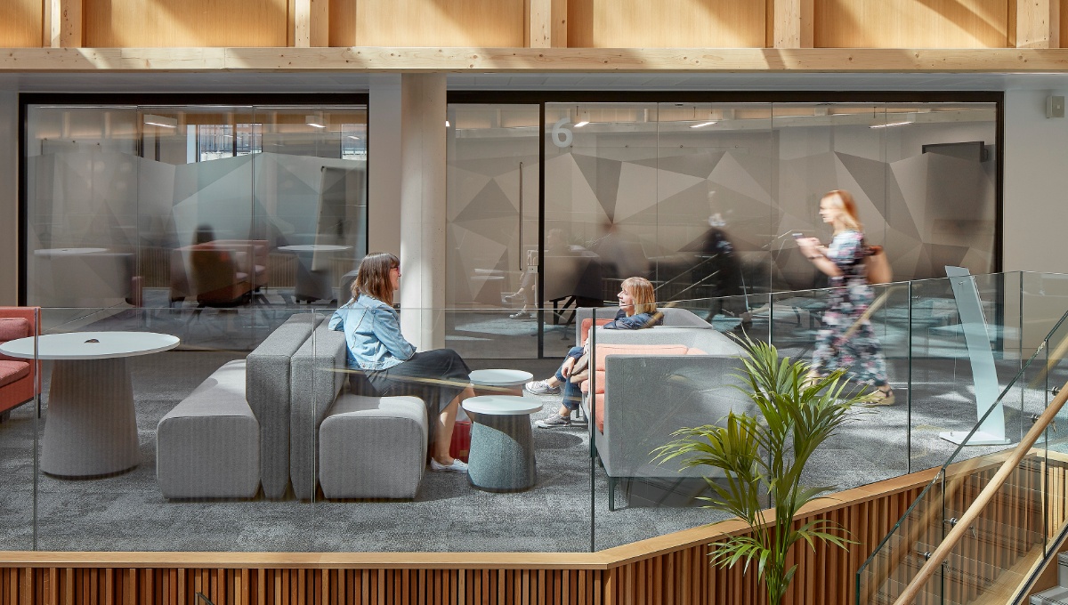 Image of two women sitting opposite each other on sofa's conversing, a woman walks past in the background