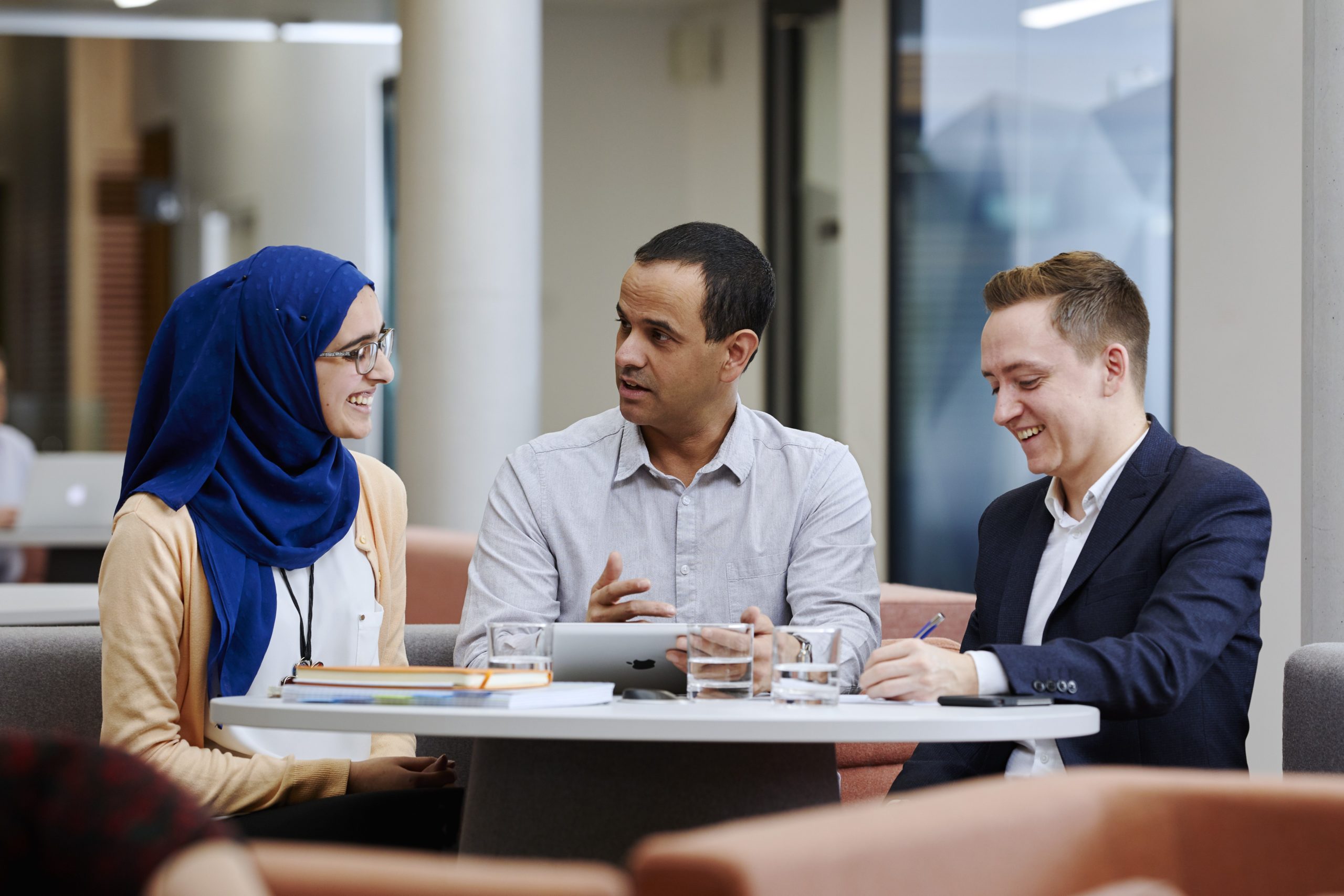 Female wearing a headscarf talks to two males sat around a table in the collaboration space at Nexus