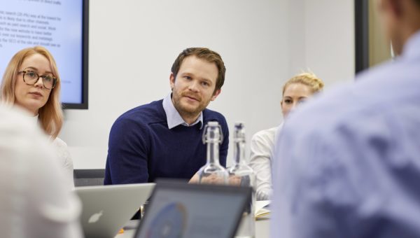 Male in centre of image talking during business meeting, female colleagues sit to left and right of him with laptops in front of them