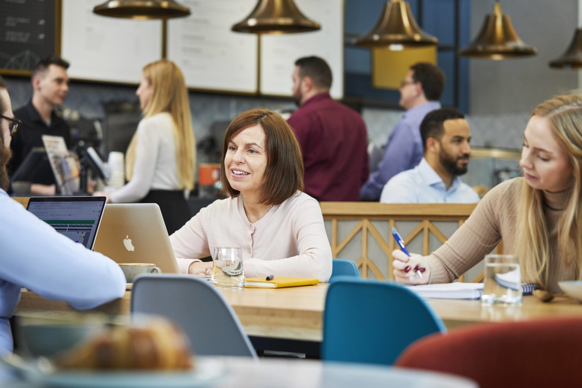Female sitting at table in a busy Nexus cafe, she is talking to a male associate as they have a coffee together