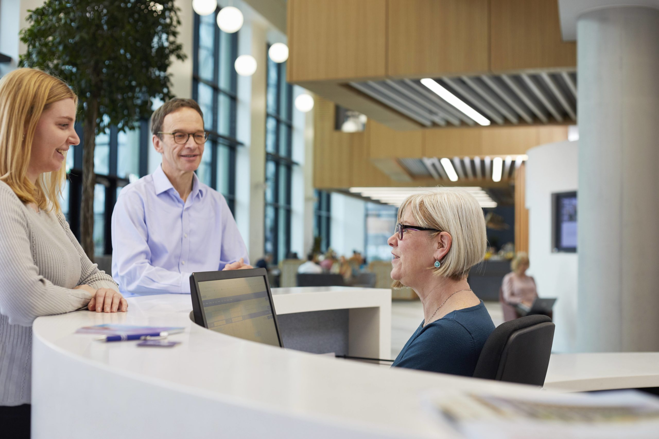 A man and woman, standing, chat to a female receptionist seated behind the Nexus reception desk