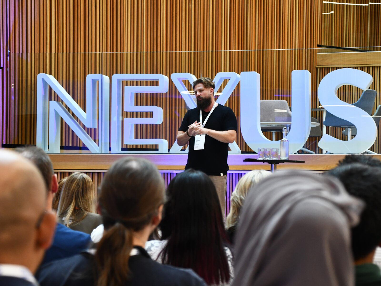 A man on a stage presenting to a crowd at an event in Nexus. There are some large letters spelling NEXUS behind him