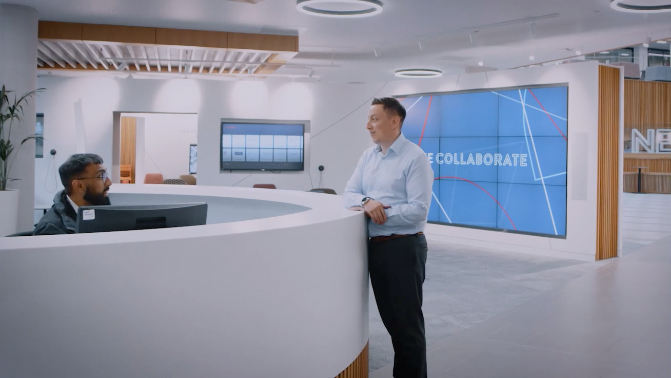 Image shows receptionist and Nexus member talking over reception desk with a live screen in the background.